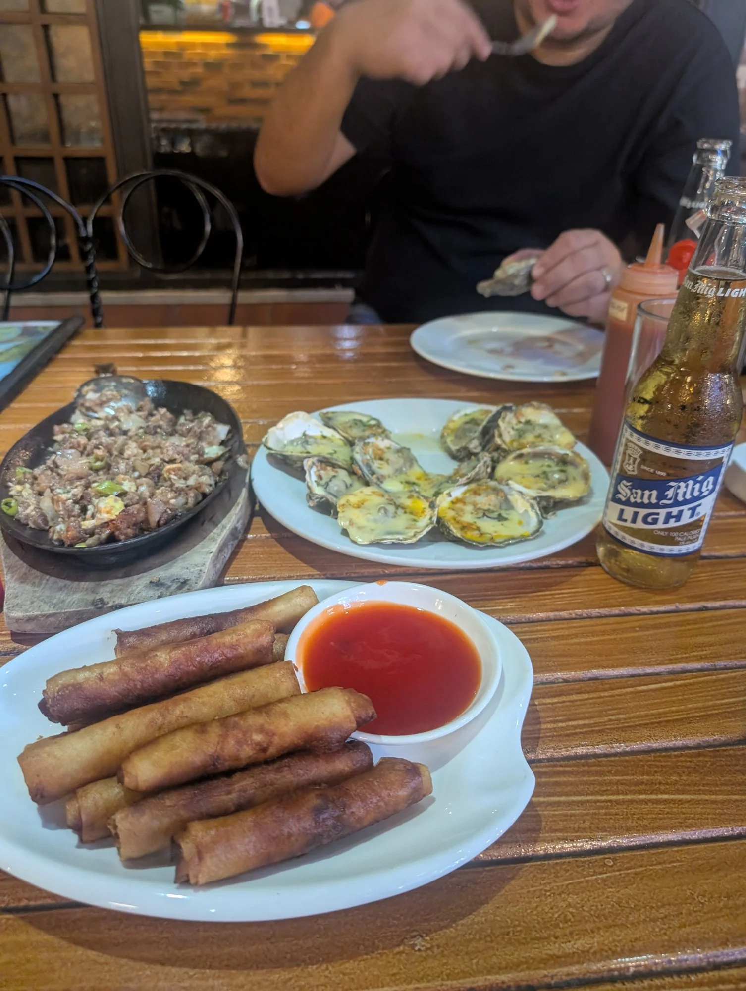 A bottle of San Mig Light beside plates of lumpia, sisig and baked oysters on a wooden restaurant table.