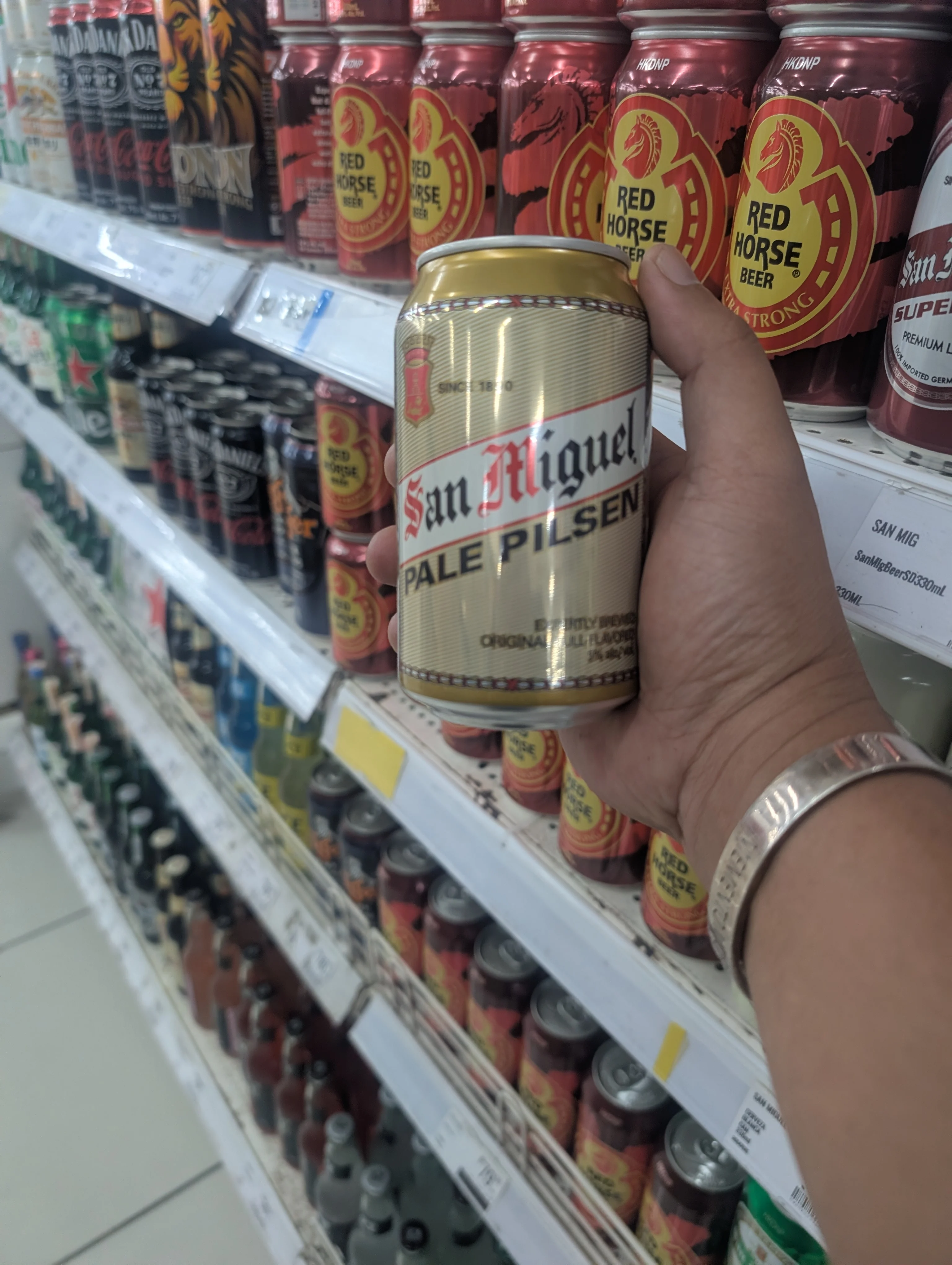 A hand holding a can of San Miguel Pale Pilsen in front of a store shelf.
