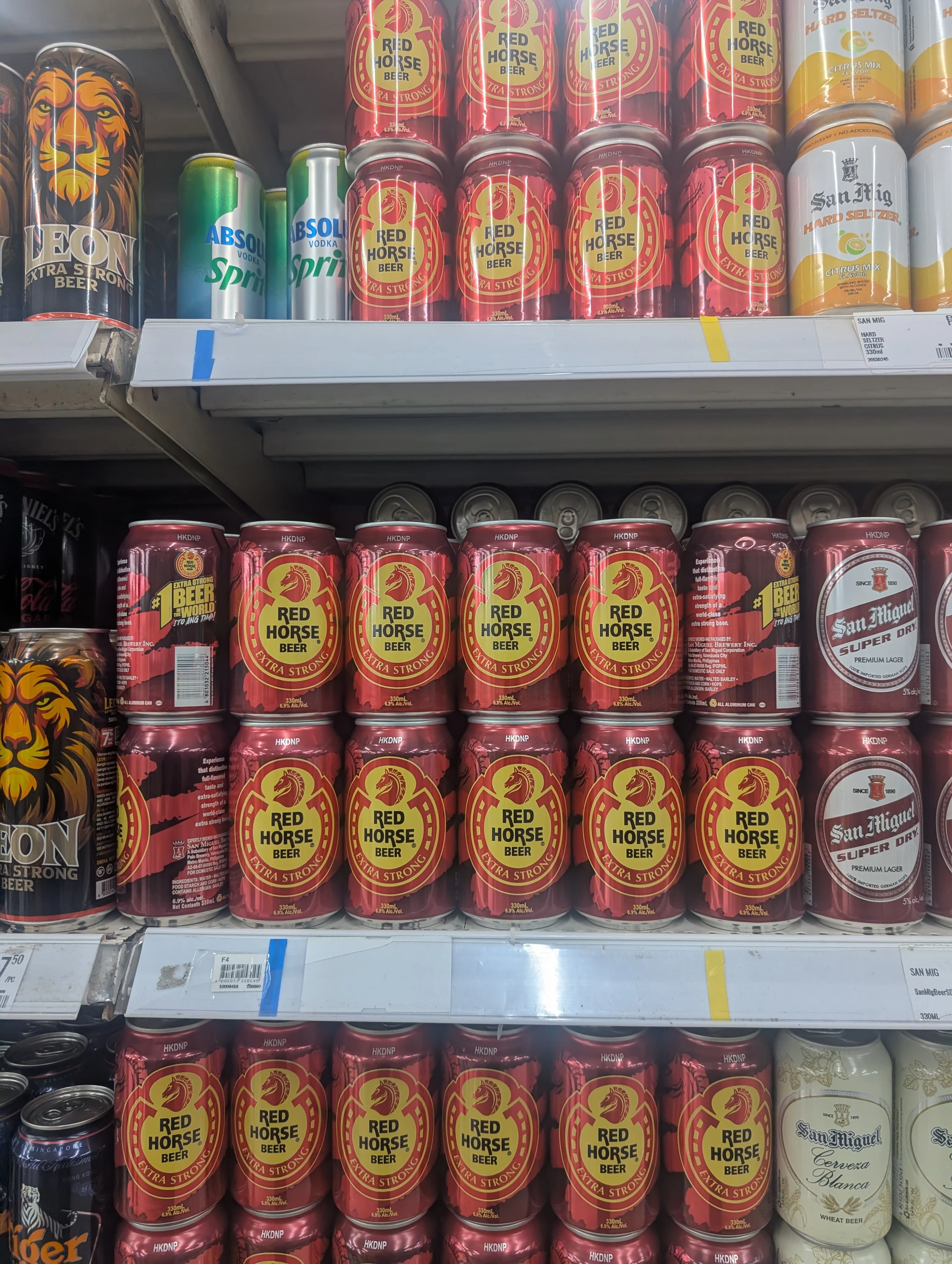 A store cooler shelf stacked with cans of San Miguel Pale Pilsen, Red Horse, San Mig Light and imported beers.